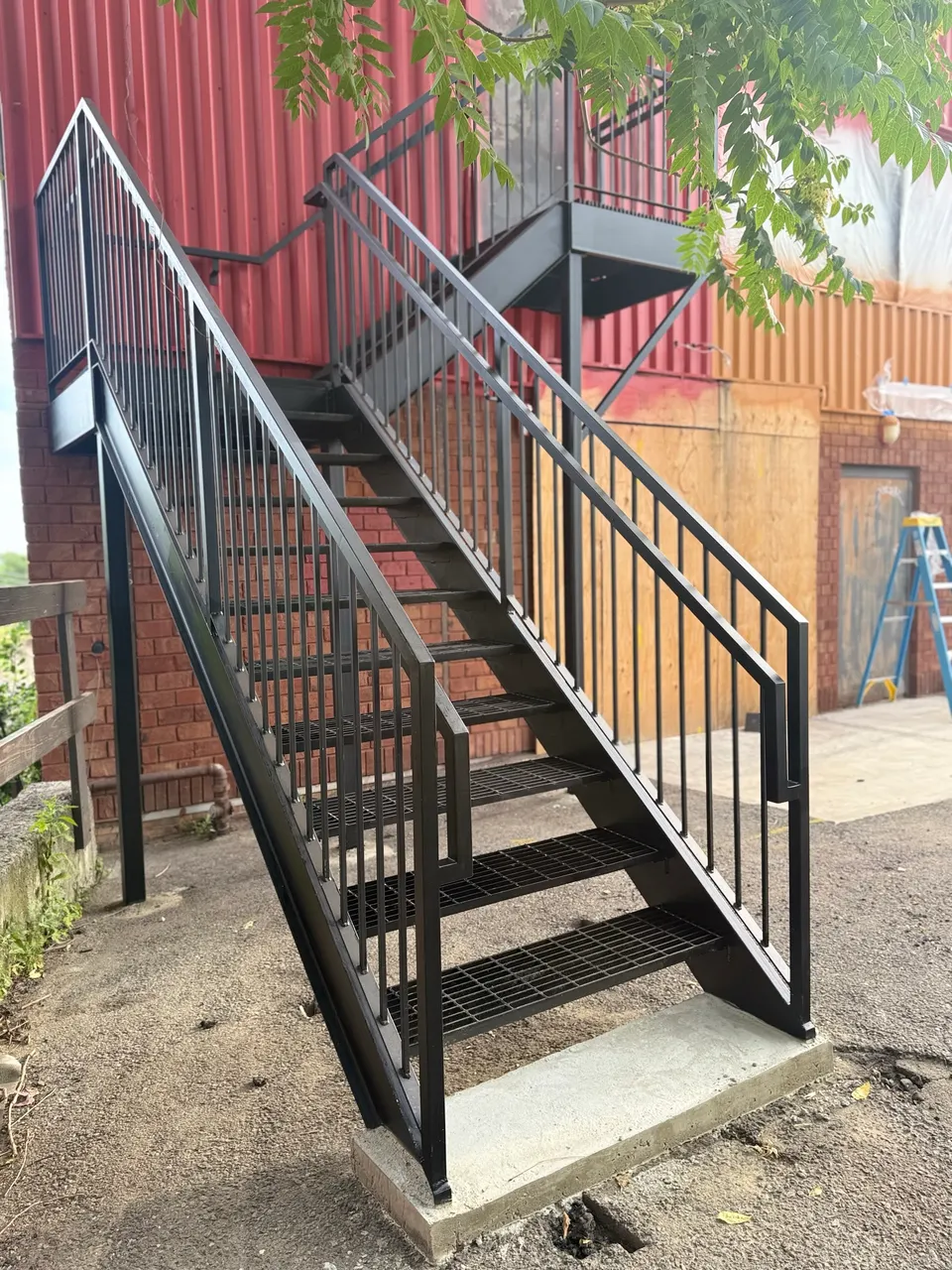 Black steel exterior stair with railings and grated treads beside a red wall